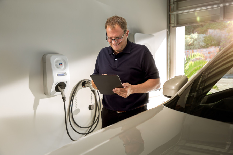 Ein mittelalter Mann mit Brille und blauem Polo-Hemd steht in einer geöffneten, weiß gestrichenen Garage. Im Vordergrund sieht man ein weißes Auto, hinter dem Mann an der Wand hängt eine Wallbox von SMA, der E-Charger. Der Mann schaut auf ein Tablet in seiner Hand.